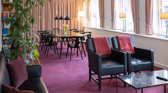 Ett modernt rum med en magentafärgad matta har ett långt bord omgivet av svarta stolar. Svarta lädersoffor med röda kuddar vetter mot ett soffbord i marmor. Fönster från golv till tak har mjukt rosa gardiner, och det finns krukväxter och hyllor i bakgrunden.